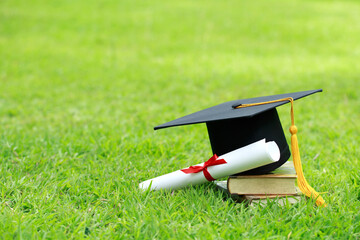 A mortarboard and graduation scroll, tied with red ribbon, on a stack of books on green grass...