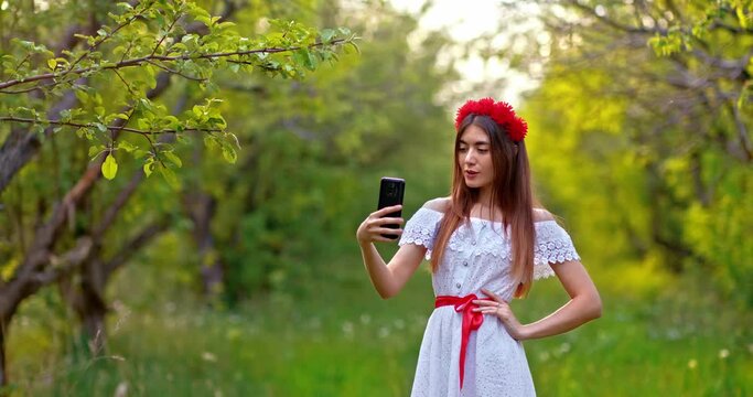 Beautiful young woman in a white dress is photographed in the park, makes a selfie on a mobile phone. Woman with long hair posing and looking into the smartphone camera, taking a selfie photo. 4k