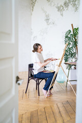 Painter in a workshop. View from the door. Young female artist draws sitting in front of easel