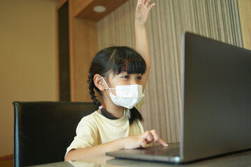 Asian kid girl school pupil wearing protective medical mask studying online from home watching web class lesson or listening tutor by video call. 