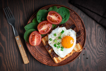 Top view of american breakfast consisting of fried eggs with yellow yolk dressed with onion on grilled toast bread with bacon, tomatoes and spinach served on plate with fork for breakfast. Horizontal