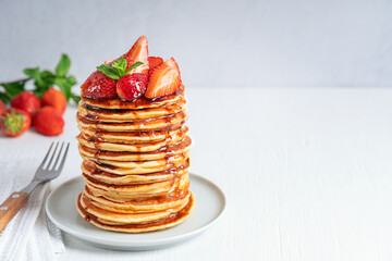 Pile of homemade pancakes or crepes decorated with strawberries, mint leaves and syrup topping served on gray round plate on white wooden table with fork and towel. Horizontal image with copy space