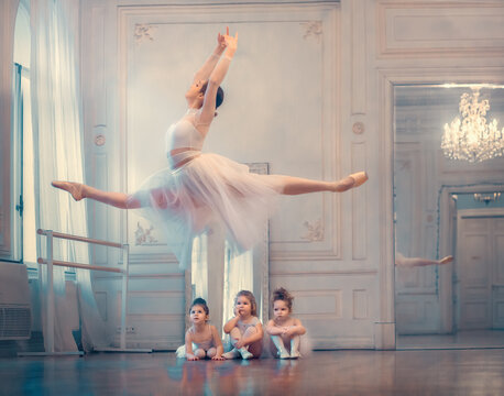 Fototapeta Three little funny ballerinas are looking with delight at gorgeous jump of adult ballerina in ballet class. Image with selective focus and toning
