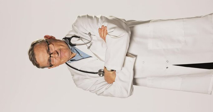 Mature doctor, dressed in a white gown, stethoscope around his neck, glasses, proudly stands and crosses his arms over his chest, looks into the camera, confident, professional