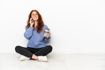 Fototapeta na wymiar Teenager redhead girl sitting on the floor isolated on white background holding coffee to take away and a mobile