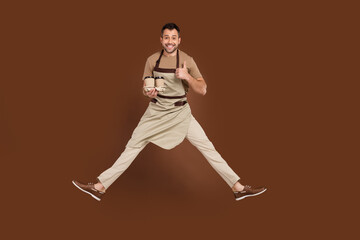 Fototapeta na wymiar Photo of confident sweet young guy dressed beige uniform jumping showing thumb up preparing latte isolated brown color background