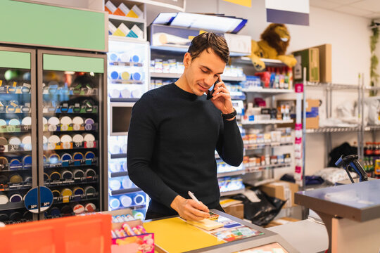 Butiks&auml;gare pratar i telefon och skriver p&aring; lapp st&aring;ende bakom kassa