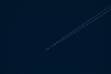 A white passenger plane flies in the distance through a dark blue sky, leaving behind a trail of...