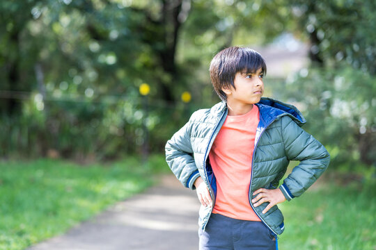 Young boy at park. Child with hands on waist wearing puffer jacket at park looking way from camera.