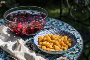 Plate with yellow raspberries and glass bowl with cherries in water on garden table.