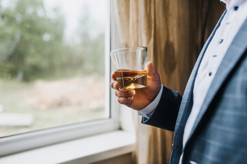 A man in a blue suit, a businessman, a director holds in his hand a glass of whiskey, cognac, rum against the background of the window. Photography, concept.