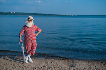 An elderly gray-haired man in a classic bathing suit and hat stands on the beach with an umbrella and waves his hand in greeting. Retired with a beard on vacation.