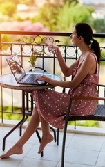 A young dark-haired woman works freelance on a hotel balcony on a sunny day. Self-isolation, work at home concept. Online work
