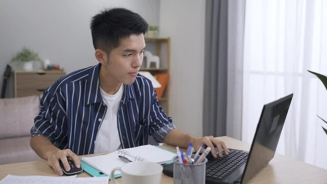 asian guy working from home is referring to the paper while typing financial report on the computer in the living room.