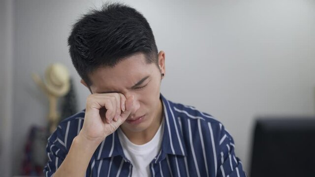 slow motion asian young man sitting behind the computer monitor is shutting and rubbing his eyes in pain while suffering unclear vision working from home.