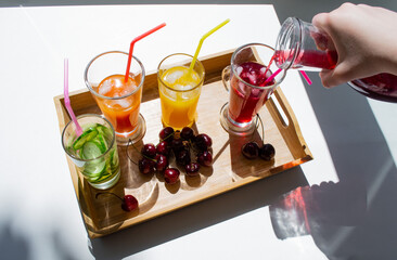 A wooden tray with colorful drinks: orange, yellow, and cucumber lemonade. Nearby are cherries. A hand pours red juice from a decanter into a glass. Contrasting shadows on a white background