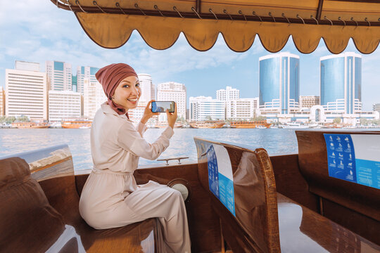 Happy asian woman in a red turban takes pictures on her smartphone while cruising on a traditional Abra Dhow boat on Dubai Creek. Travel in UAE concept