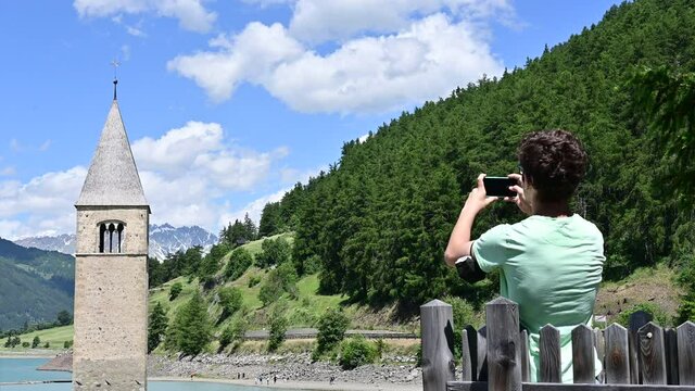 Curon, Italy, June 2021. Footage in beautiful summer day of the old bell tower of Curon (Graun) emerging from the waters of Lake Resia. A guy in a green shirt takes a picture with his smartphone.