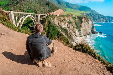 A young man in a denim jacket on a slope overlooking the Bixby Creek Bridge on the Big Sur coast in...