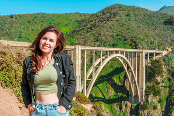 A young beautiful woman is traveling with a view of the Bixby Creek Bridge on the Big Sur coast in...