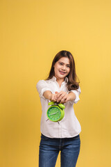 Beautiful Asian woman standing and hold a green alarm clock over yellow background smiling happily.