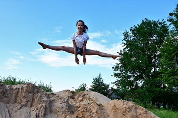 cute cheerful girl in jump does twine in air against background of blue sky. Female athlete in white T-shirt and denim shorts performs acrobatic exercises. Copy space
