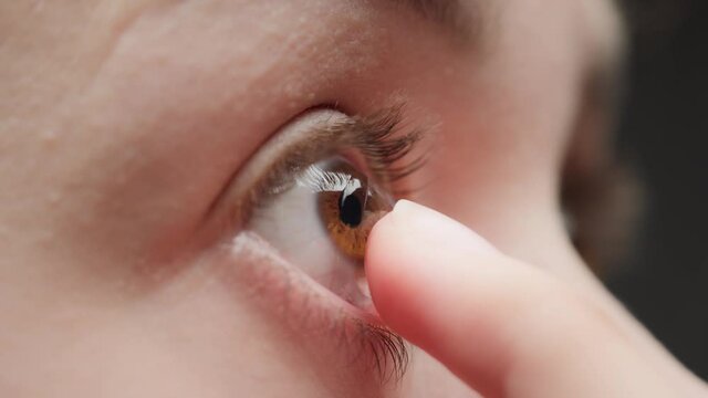 Close-up of a brown-eyed young woman putting on the contact lens with fingers and blinking. Advertising of contact lenses for vision improvement. Optical shop, health care and medicine concept.