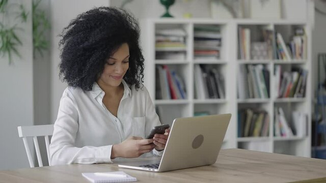 Portrait of a young African American woman texting on a smartphone. A brunette with curly hair in a white blouse sits at a table in a light home office. Close up. Slow motion ready 59.97fps.