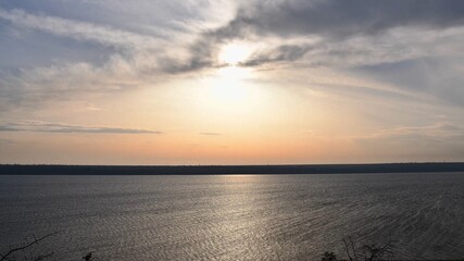 Obraz premium White sun shines in high sky with light spindrift clouds over tranquil water surface. Sunshine over shore landscape with blurry sunlight reflection