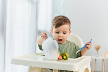 Child holding spoon and pouring vegetables on high chair