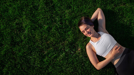 A beautiful, cheerful, cheerful woman in a white T-shirt is lying on the green lawn in the park and...