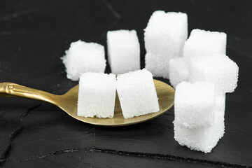 Several white sugar cubes were placed in a brass spoon on a black stone background.