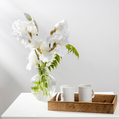 A bouquet of three white irises and fern in a transparent vase on the table. Two ceramic tea in the wooden tray. Breakfast