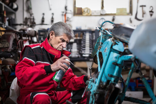 Elderly mechanic spraying oil on motorcycle detail in workshop