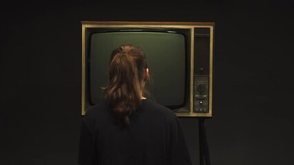 Young woman turning on old retro television on black background, searching for a signal, channel setting, pressing a button on old-fashioned TV, bad signal reception, cinematography concept.