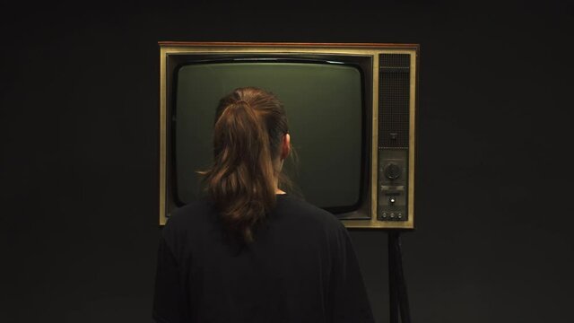 Young woman turning on old retro television on black background, searching for a signal, channel setting, pressing a button on old-fashioned TV, bad signal reception, cinematography concept.
