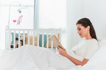 Side view of woman reading book on bed near blurred baby crib