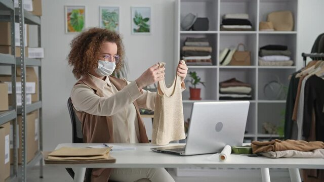work in picture, young female entrepreneur wearing protective mask takes an order in an online store uses laptop while working in quarantine, smiles and looks at camera