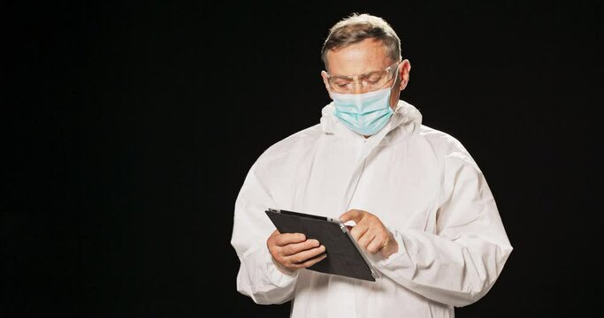 An elderly men dressed in protective clothing, white gown, mask and safety glasses, searches for information on a tablet, checks the results of virus tests on isolated patients. Black background