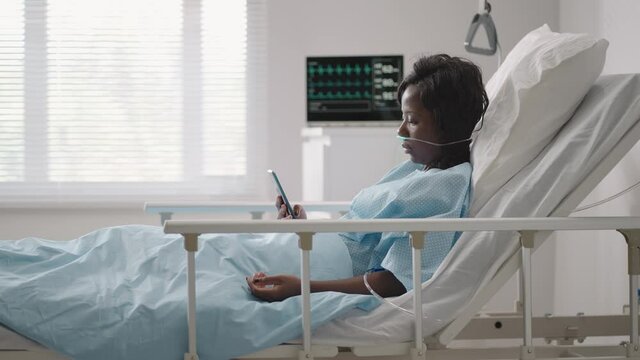 Black Woman Patient In Hospital With Saline Solution Volumetric Infusion Pump Using Mobile Phone On Examination Couch. African Women Lying In Hospital Bed With Smart Mobile Phone While In Hospital.