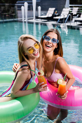 excited woman in sunglasses and swimsuits relaxing with cocktails in pool