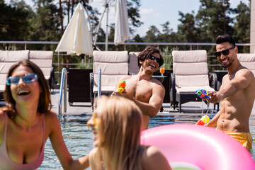 cheerful interracial men playing with water guns near blurred women in swimming pool
