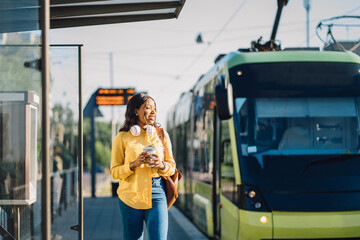 Attractive african american female traveler or student waiting for public transport standing on bus stop outdoor at sunny morning with transport on blurred background. © Iryna