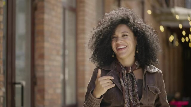 Portrait of young African American woman waving hand, showing gesture come here. Brunette in brown leather jacket posing on street against blurred brick building. Close up. Slow motion ready 59.97fps.