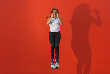 Latin, redhead woman in sportswear jumping with closed arms and legs wearing Kangoo Jumps boots on orange background. Healthy life, activity and sport concept