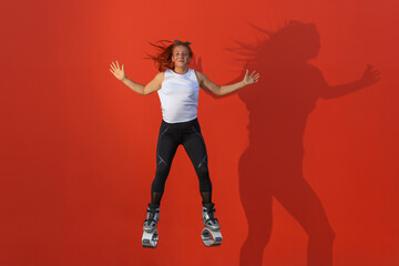 Fototapeta na wymiar Latin, redhead woman in sportswear jumping with open arms wearing Kangoo Jumps boots on orange background. Active movement, action, fitness and wellness concept
