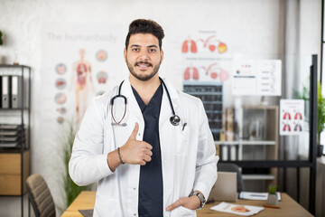 Competent young Muslim Arabian male medical worker in white lab coat showing thumb up while posing...