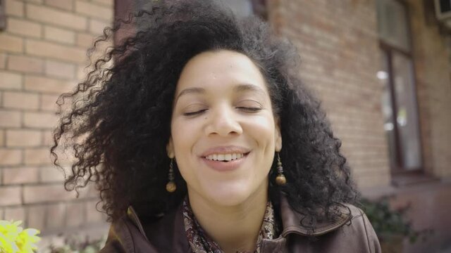 Portrait of young African American woman talking on video call. Brunette in brown leather jacket posing on street against blurred building. View from web camera. Close up. Slow motion ready 59.97fps.