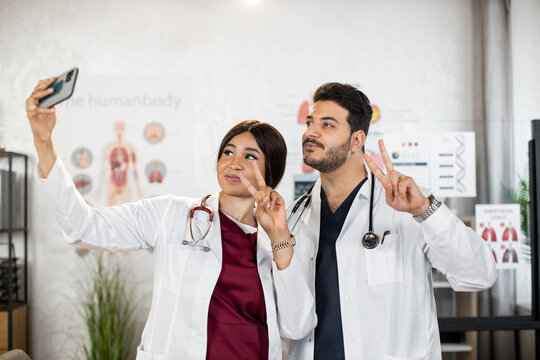 Smiling african woman and indian man in white lab coat showing sign peace with fingers while taking selfie on smartphone. Two medical workers making photo after international meeting.