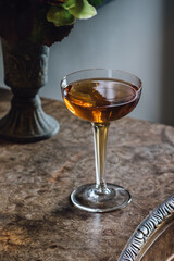 whiskey cocktail in hollow stem glass on burl wood table with vase of flowers 